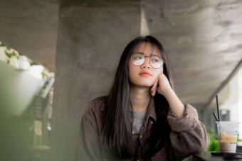 A contemplative young woman in eyeglasses, sitting indoors with a casual drink.