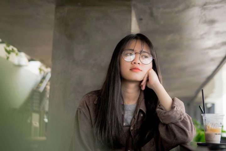 A contemplative young woman in eyeglasses, sitting indoors with a casual drink.