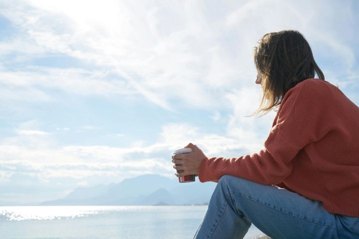 A woman in casual attire enjoys a hot drink while gazing at the sea, epitomizing relaxation.