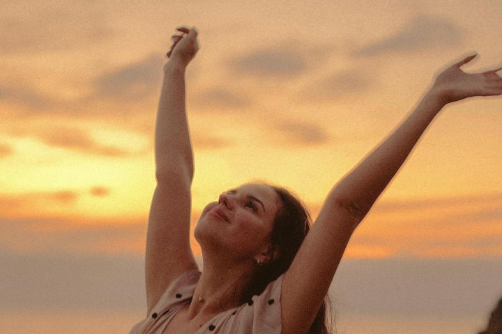 A woman in a polka dot dress expresses freedom and joy by the sea at sunset.