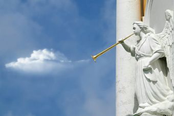 Sculpture of an angel with a trumpet, set against a bright blue sky with clouds.