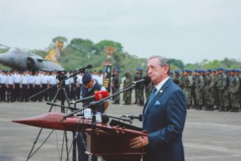Side view of mature colonel with order standing near microphones on asphalt pavement behind army of soldiers near airplane monument under serene sky during national military holiday and looking away