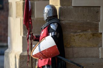A medieval knight in armor stands guard with a shield and flag outside a stone building.