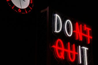 Neon sign reading 'Don't Quit' beside a wall clock at midnight. Encouragement theme in urban setting.