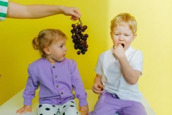 A boy enjoying grapes while sitting next to his curious sister indoors against a yellow background.