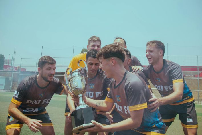 Soccer team joyfully celebrating their victory on a sunny pitch holding a trophy.