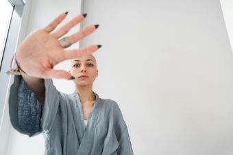Bald woman in a robe making a stop gesture indoors, symbolizing resistance or caution.