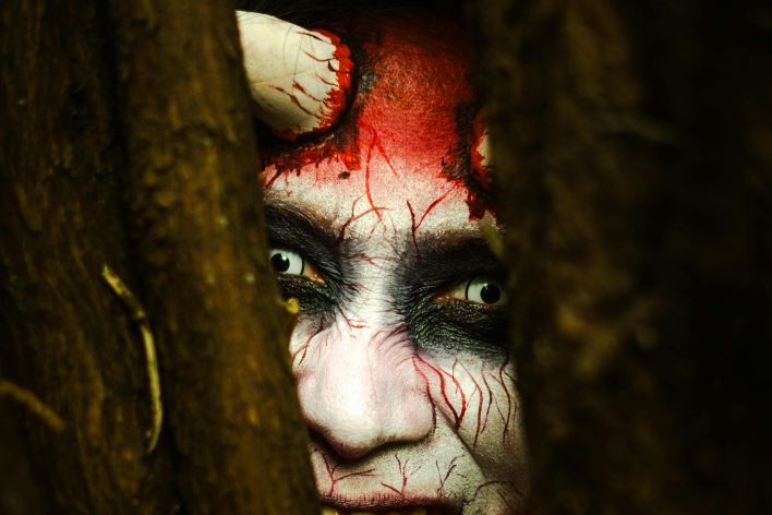 Creepy portrait of a person in demon makeup peering through trees.